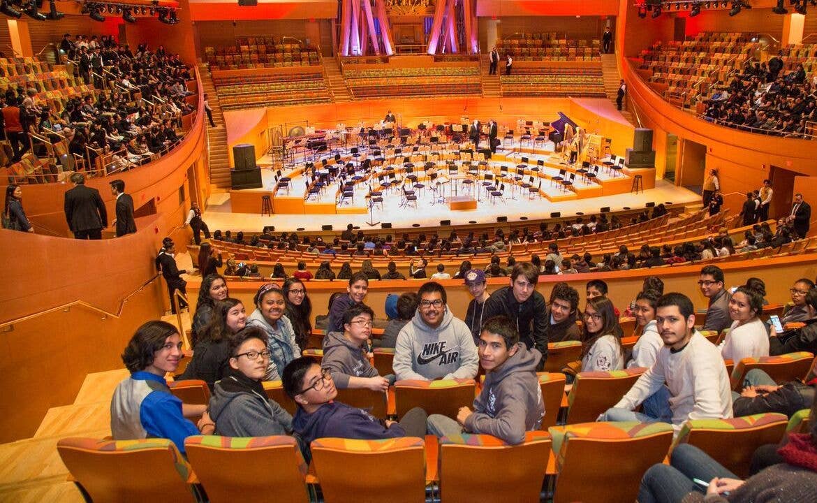 Students sit in bright auditorium seats, watching a formal session on a large stage with rows of empty desks