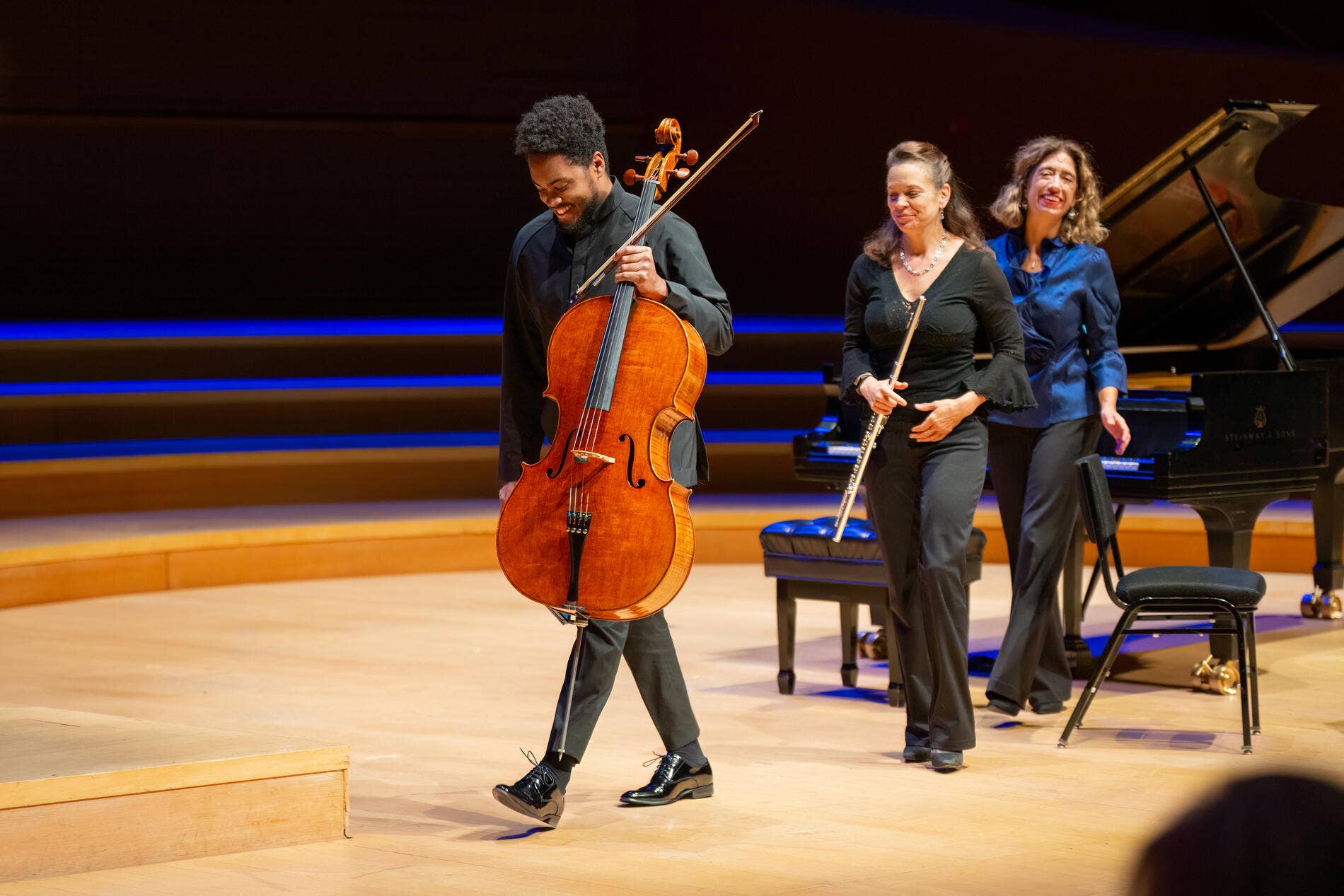 Cellist bows onstage with two smiling women beside a grand piano during a live concert performance