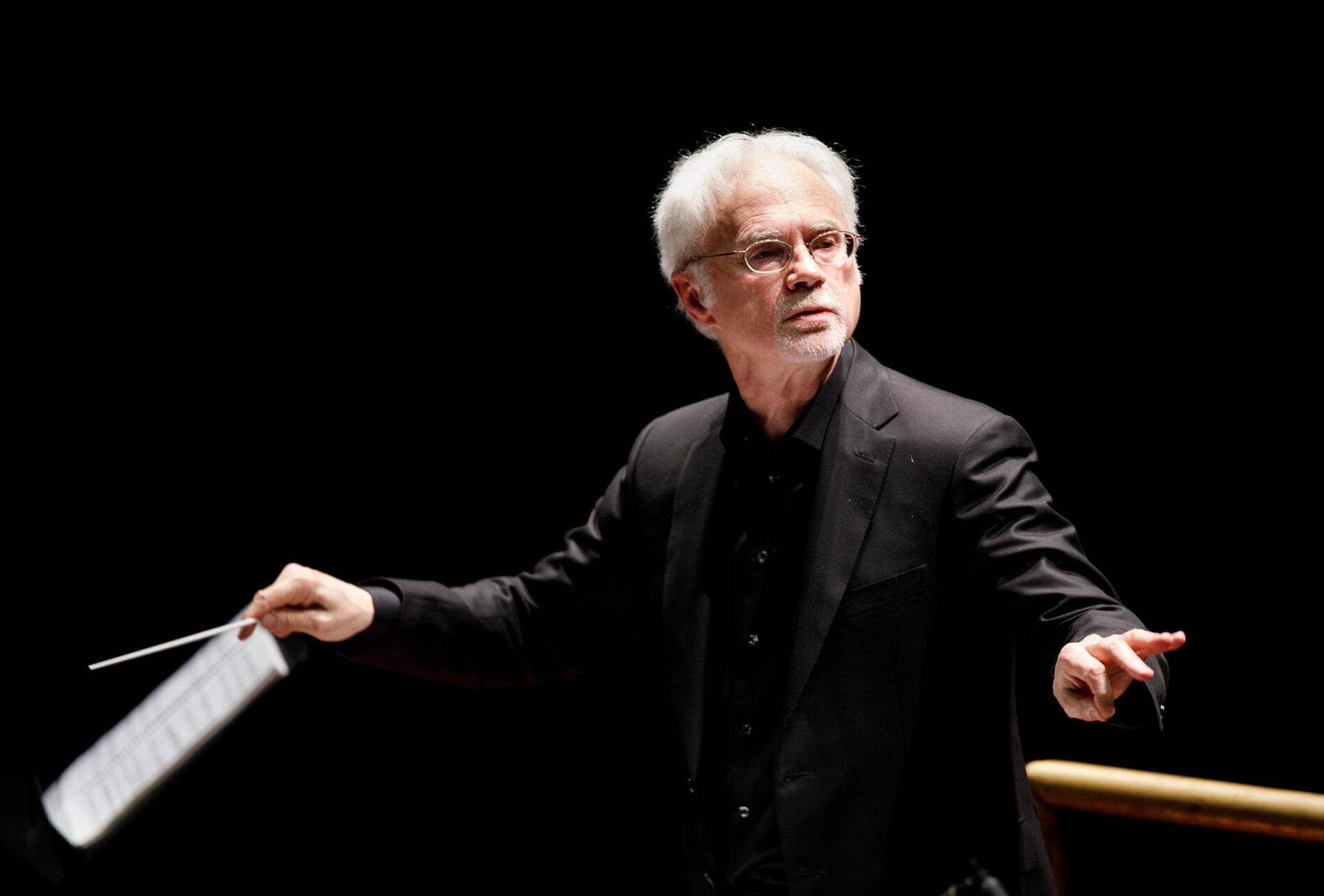 John Adams dirigiendo una orquesta en el escenario bajo una iluminación tenue.
