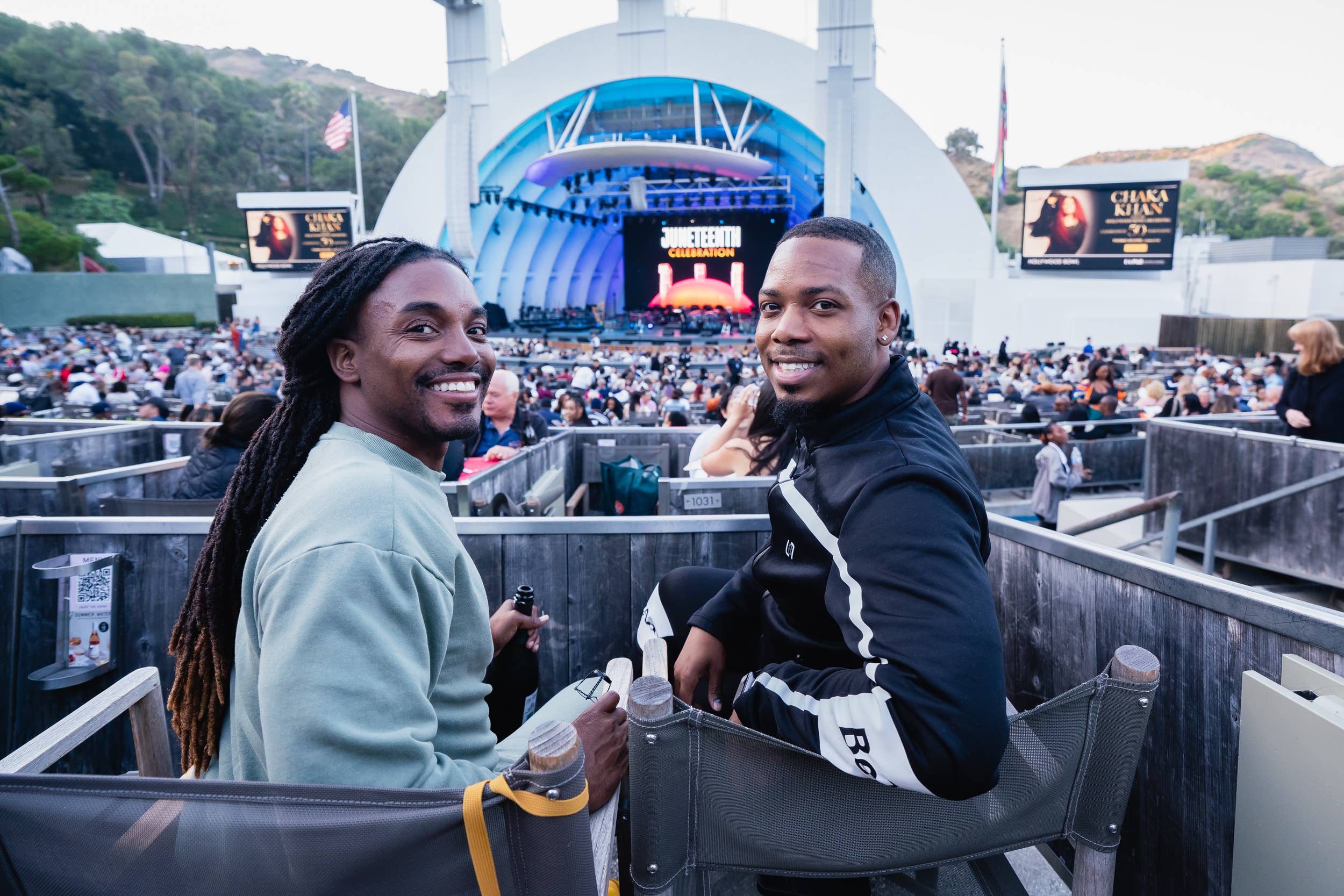 Two smiling men seated at an outdoor concert stage with a large backdrop and a cheering crowd.