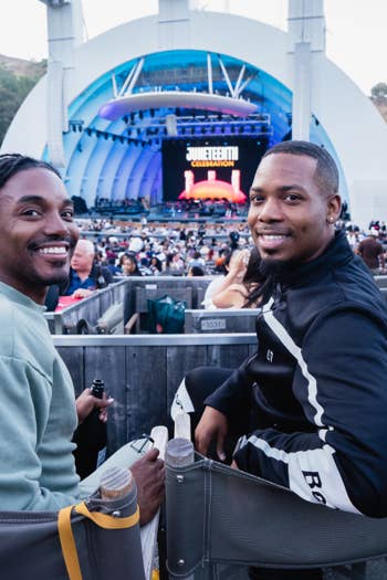 Two smiling men seated at an outdoor concert stage with a large backdrop and a cheering crowd.