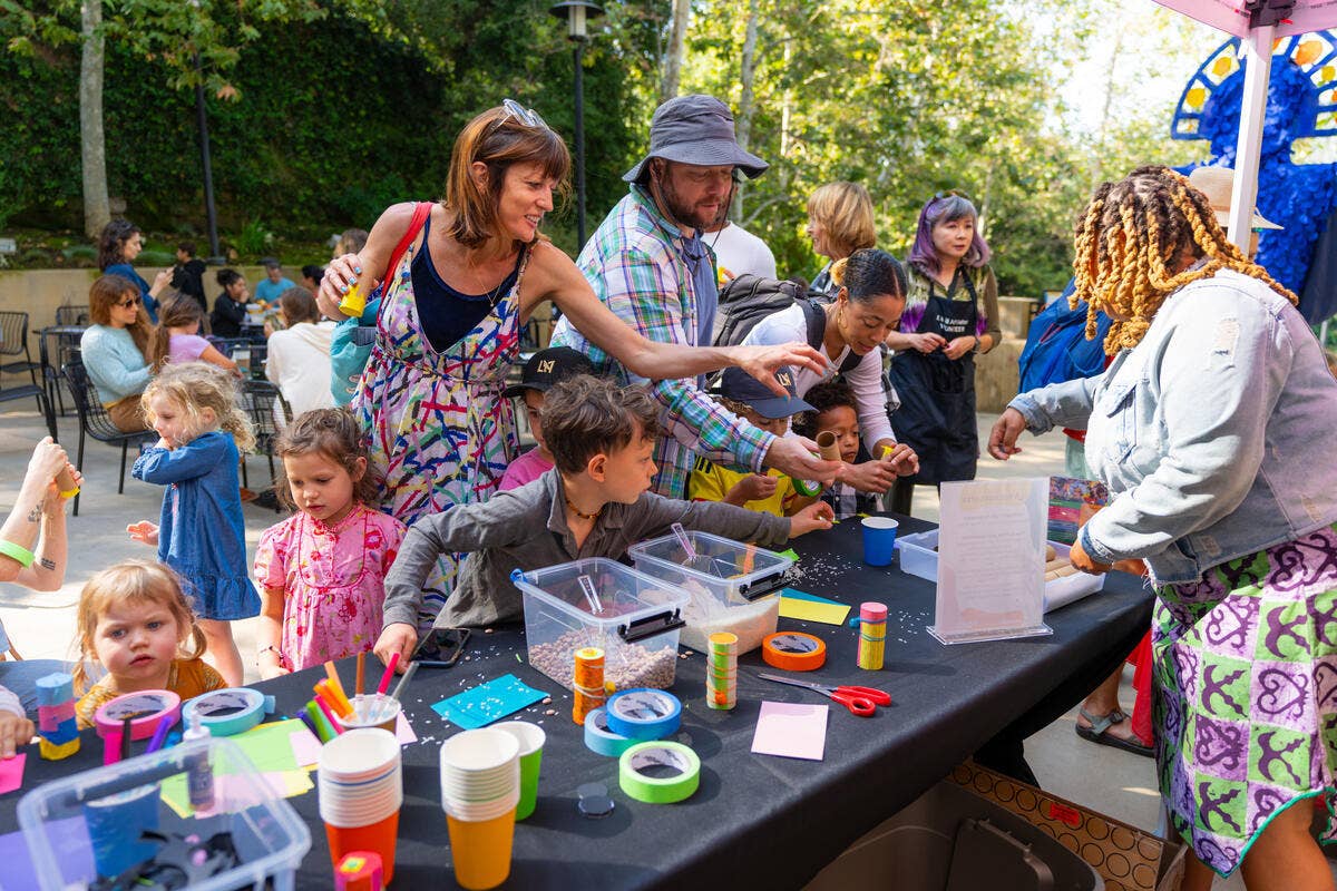 Outdoor community art table with children and adults painting, supplies and cups on a long black table.