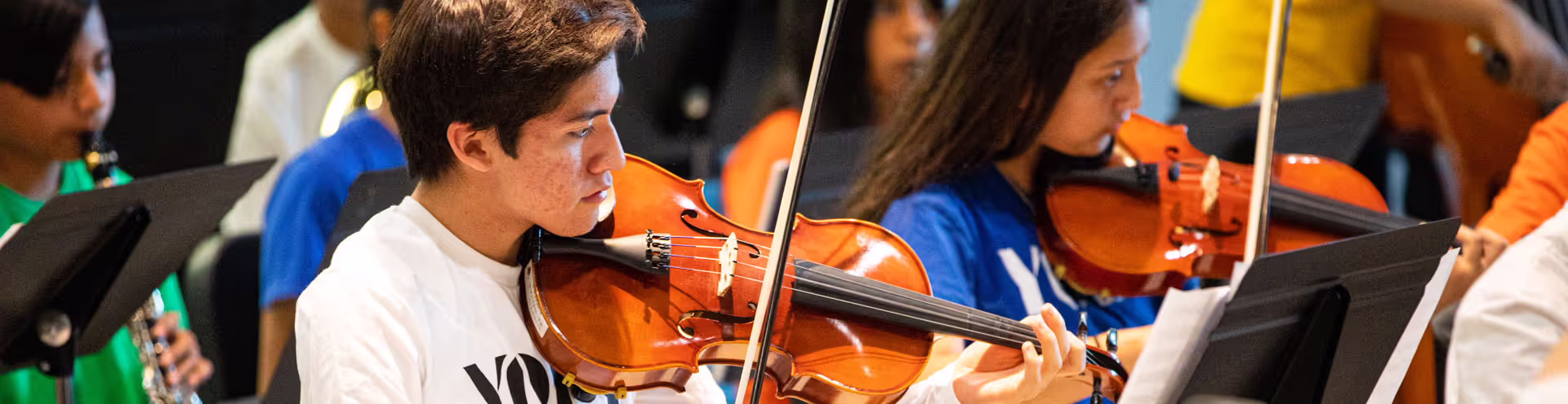 Two people pay violin with one person playing a clarinet in the background.