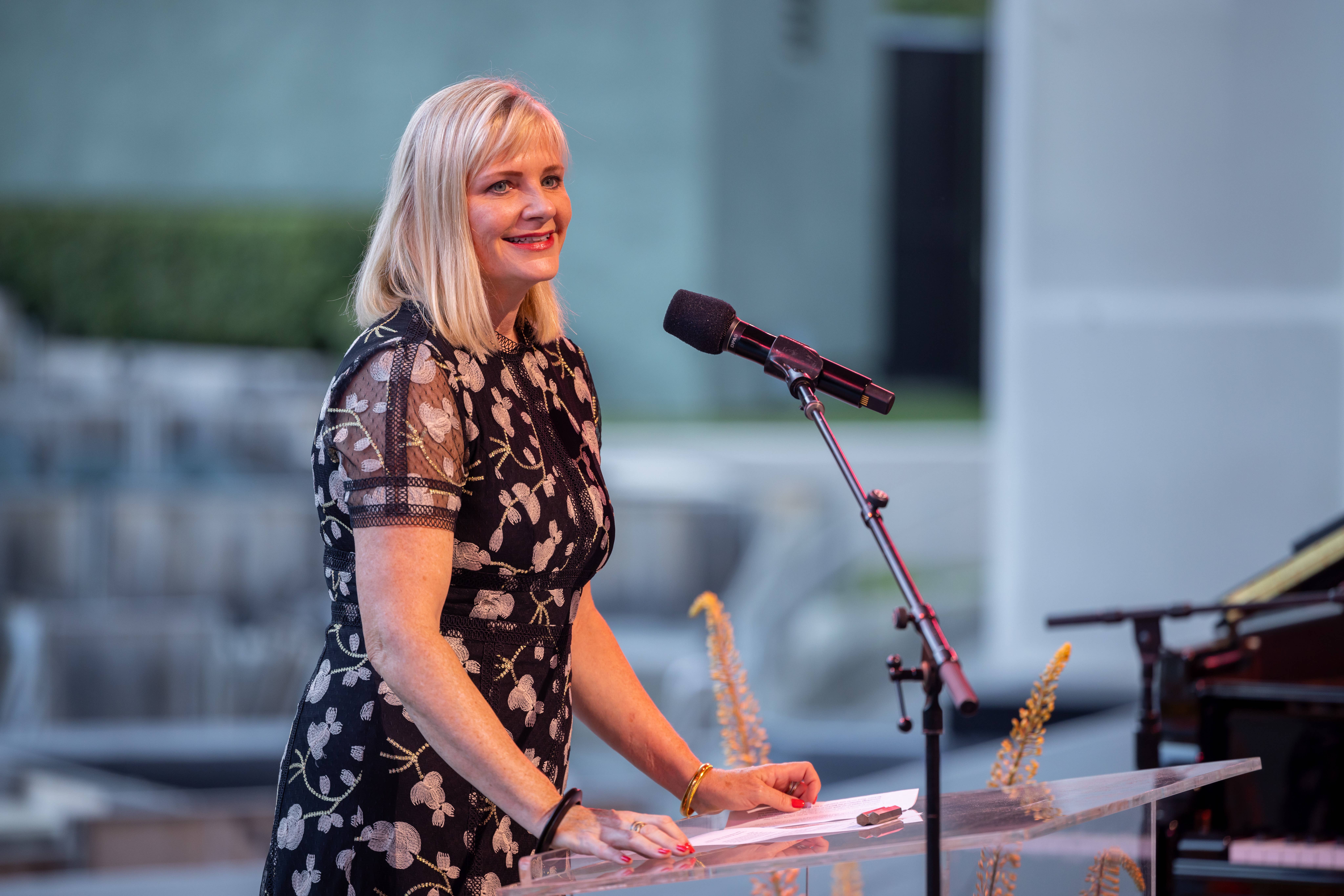 Kim Noltemy speaks at outdoor podium with microphone, wearing a patterned dress and smiling.
