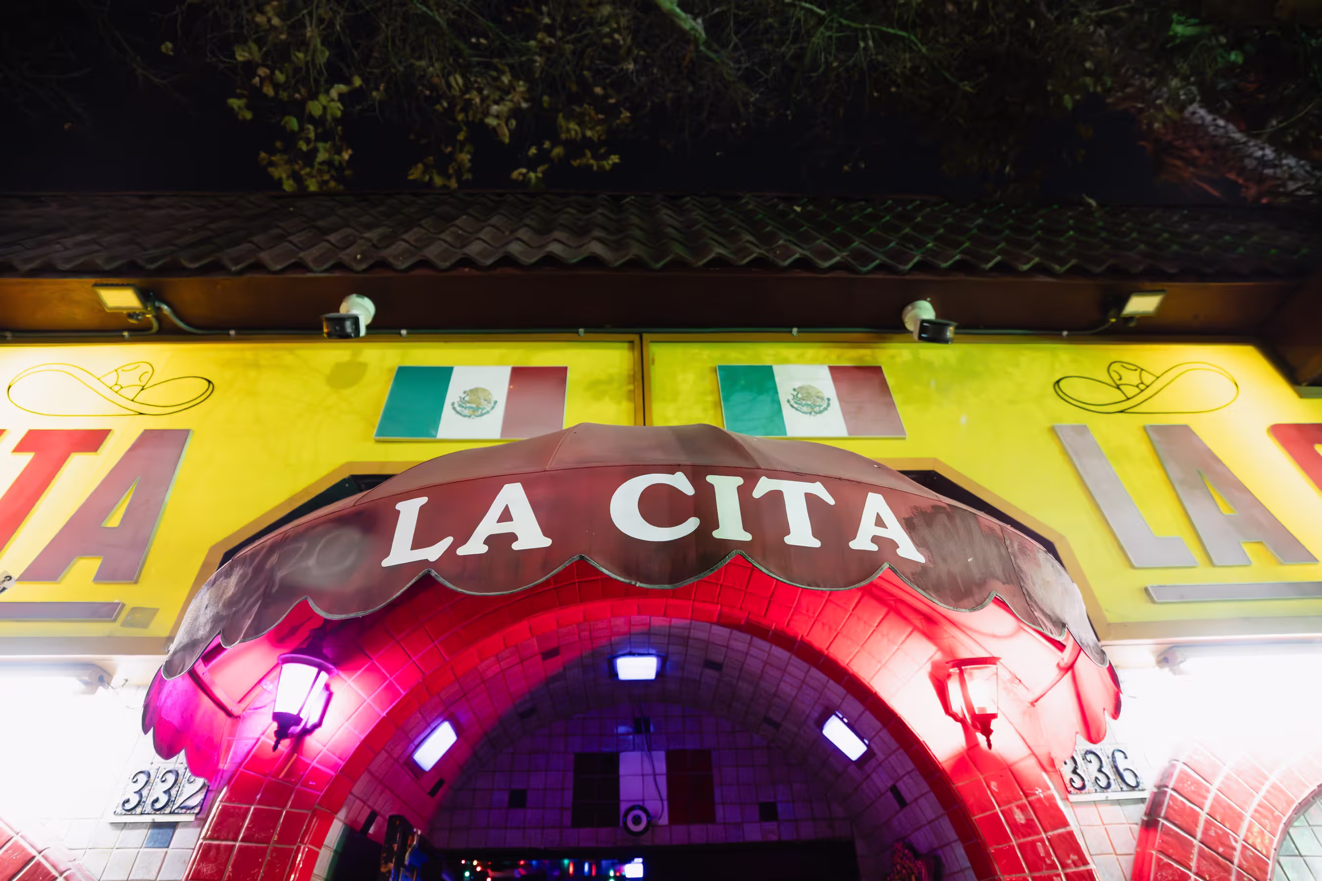 Entrance arch with LA CITA sign and Mexico City flag mural on a yellow building, under trees.