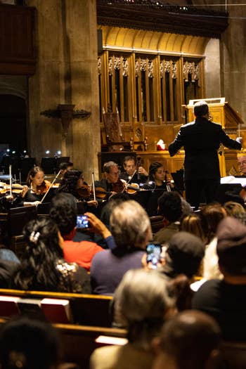 Conductor leads an orchestra inside a grand hall as musicians play string instruments under warm stage lights