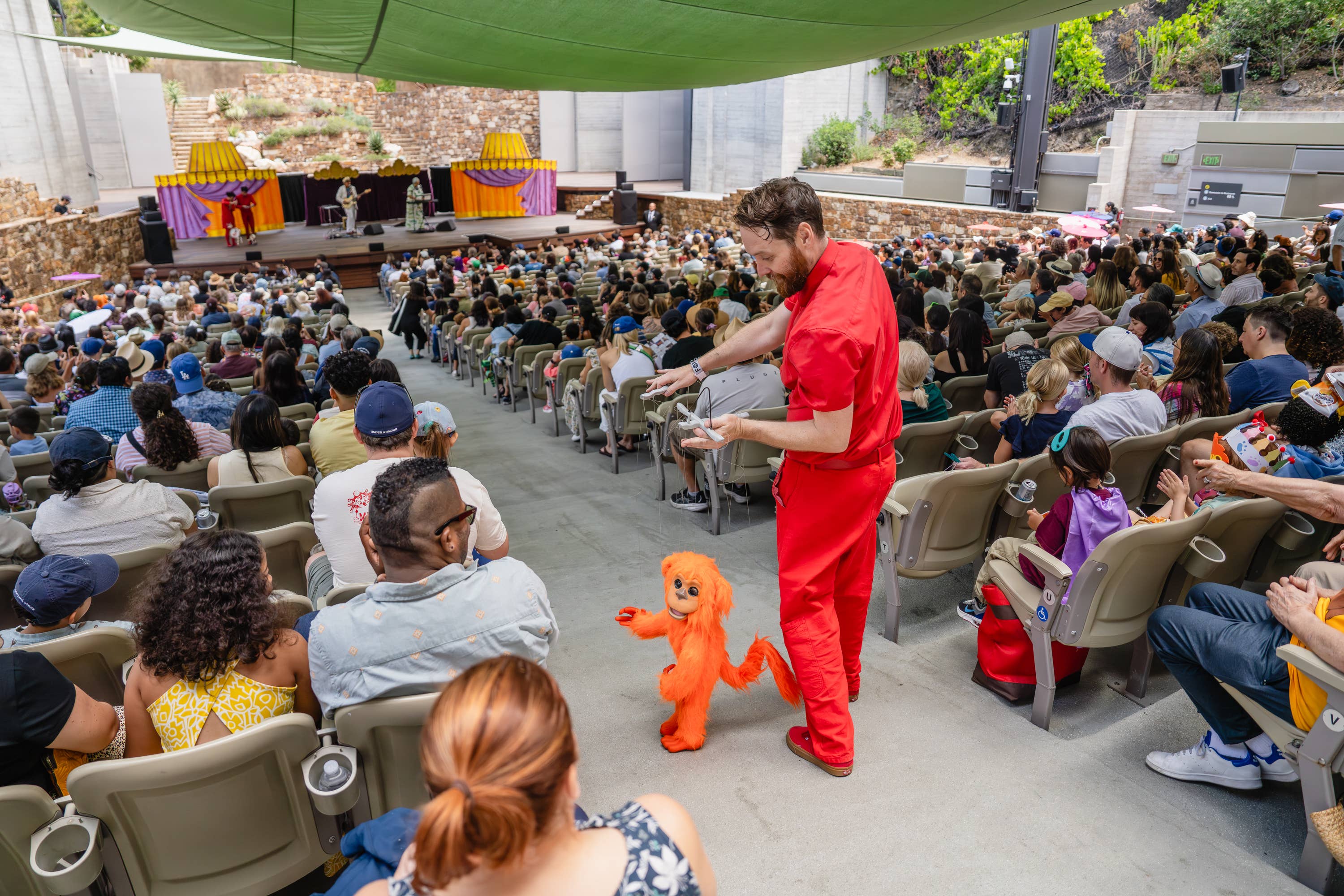 Performer in red stands on an outdoor stage aisle, entertaining a large audience with a bright orange puppet dog