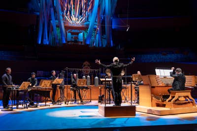 Conductor on stage with outstretched arms, orchestra seated, blue light beams and colorful backdrop in concert hall.
