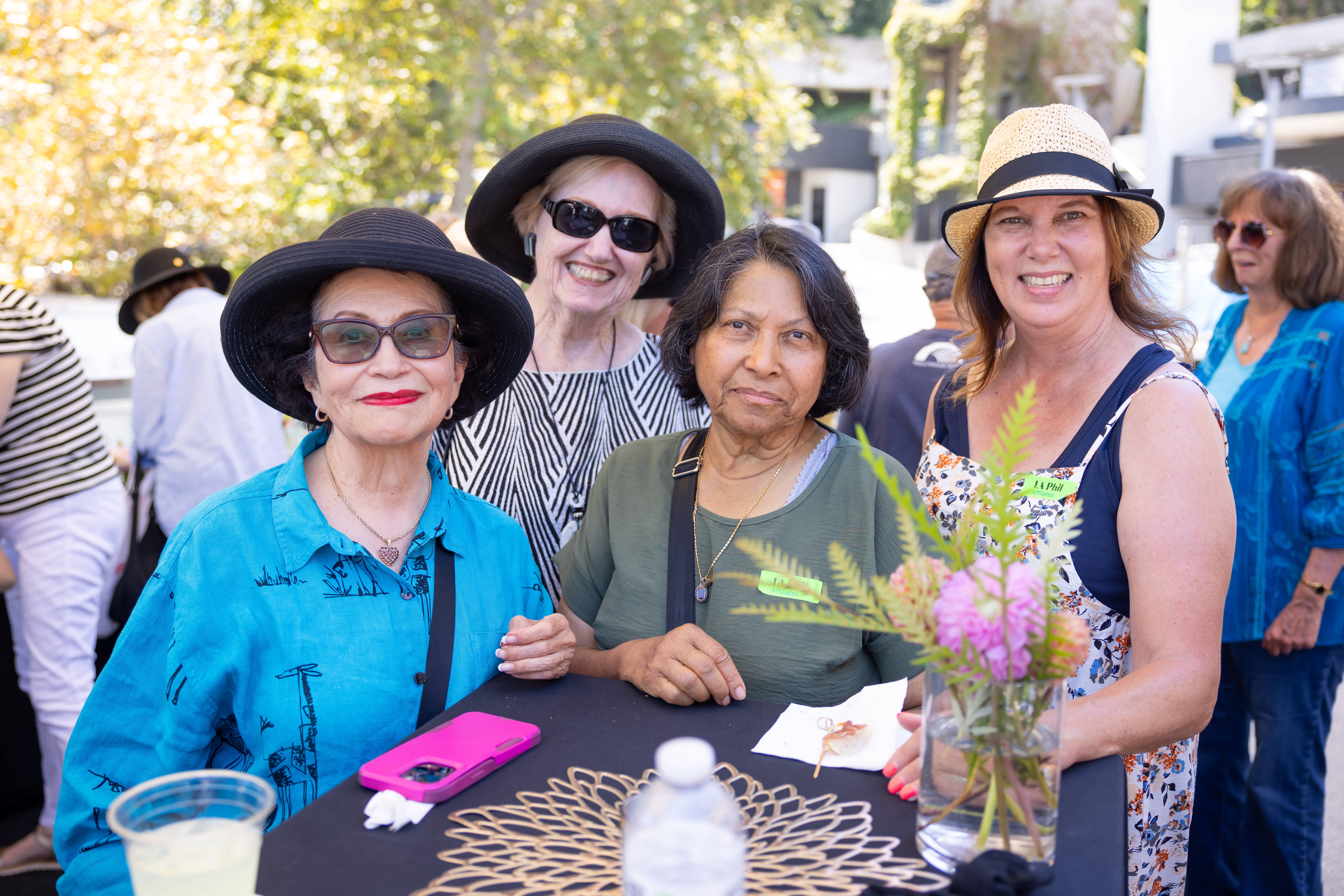 Group of women wearing hats and sunglasses smiling at an outdoor community event table with flowers and drinks.