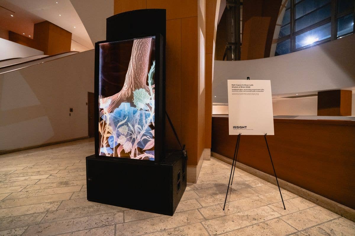 Lobby display with a lit digital art screen on a pedestal and an accompanying exhibition sign on tripods.