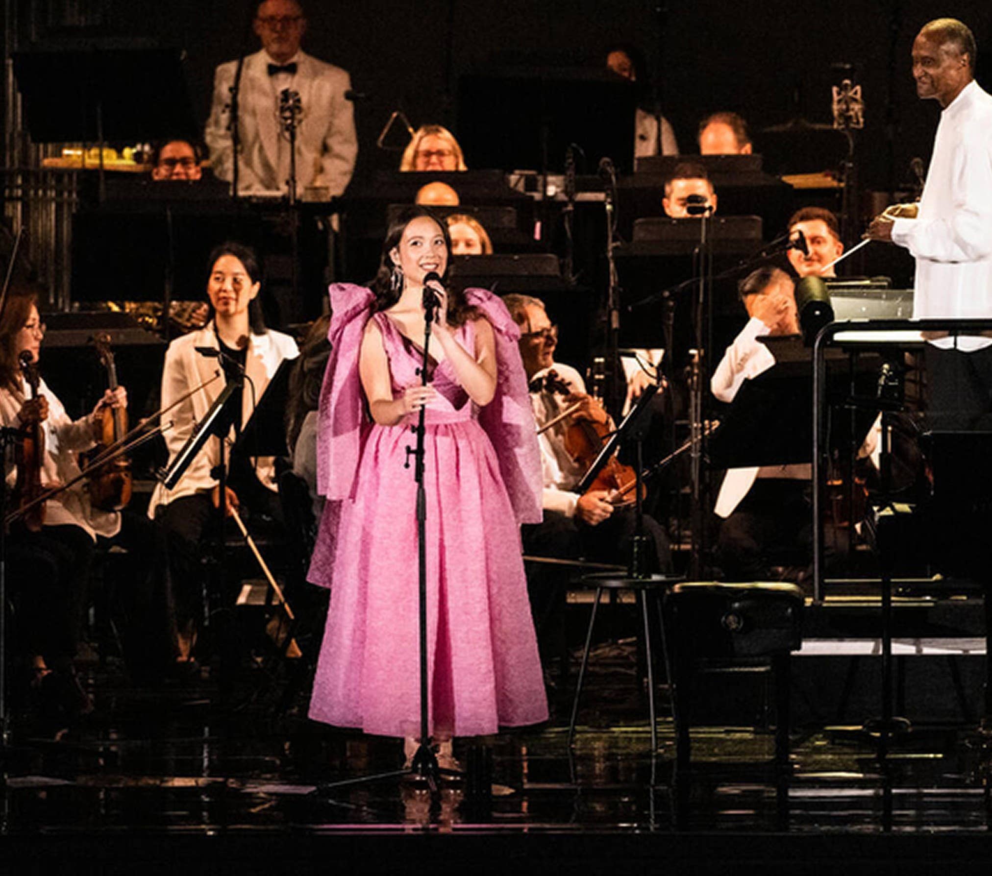 Singer in a pink gown performs onstage with a live orchestra, holding the microphone near center stage.