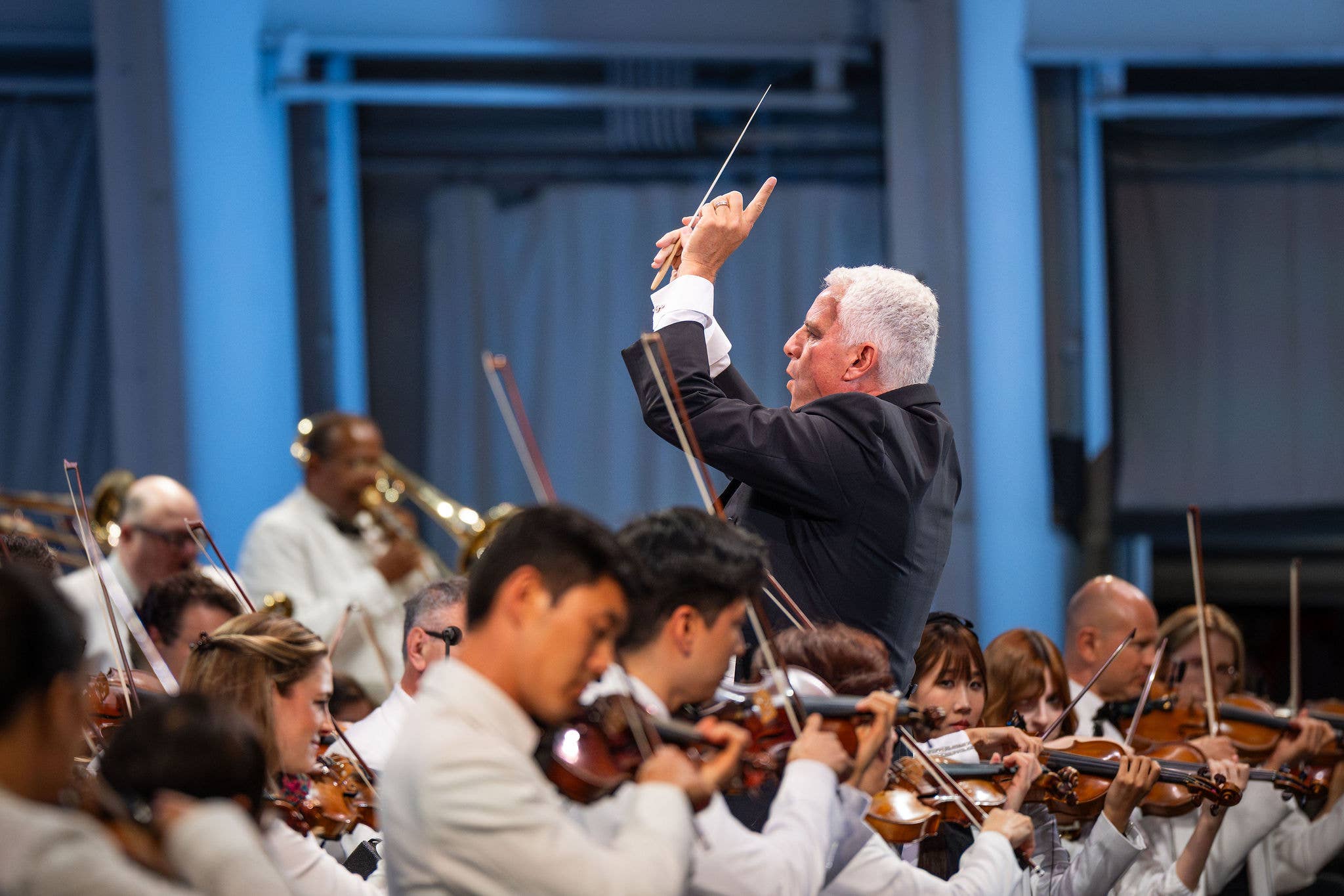 Orchestra conductor raises baton with right hand while musicians play string instruments in a concert hall.