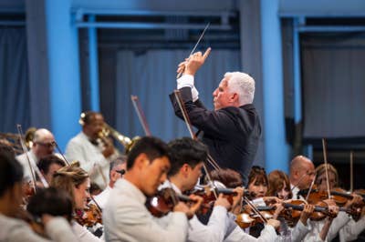 director de orquesta  batuta la mano derecha mientras los músicos tocan instrumentos de cuerda en una sala de conciertos.