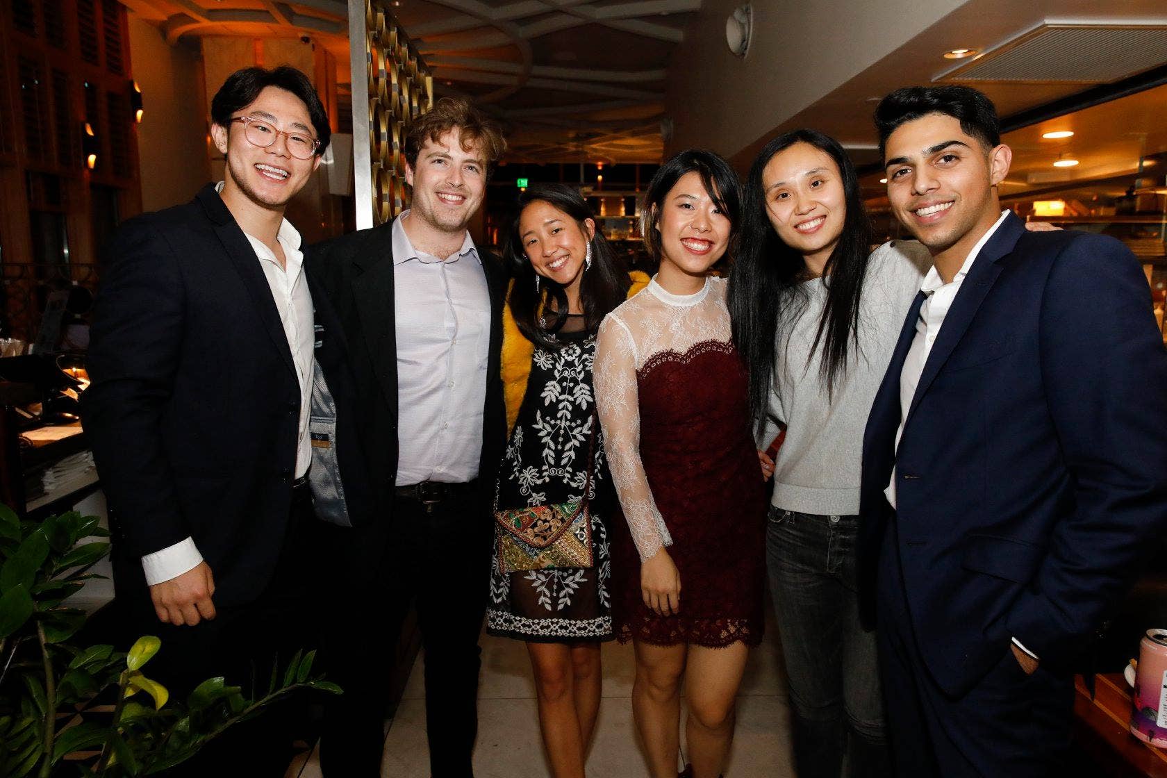 Six young adults posing and smiling in a restaurant lounge with warm lighting and bar shelves behind them