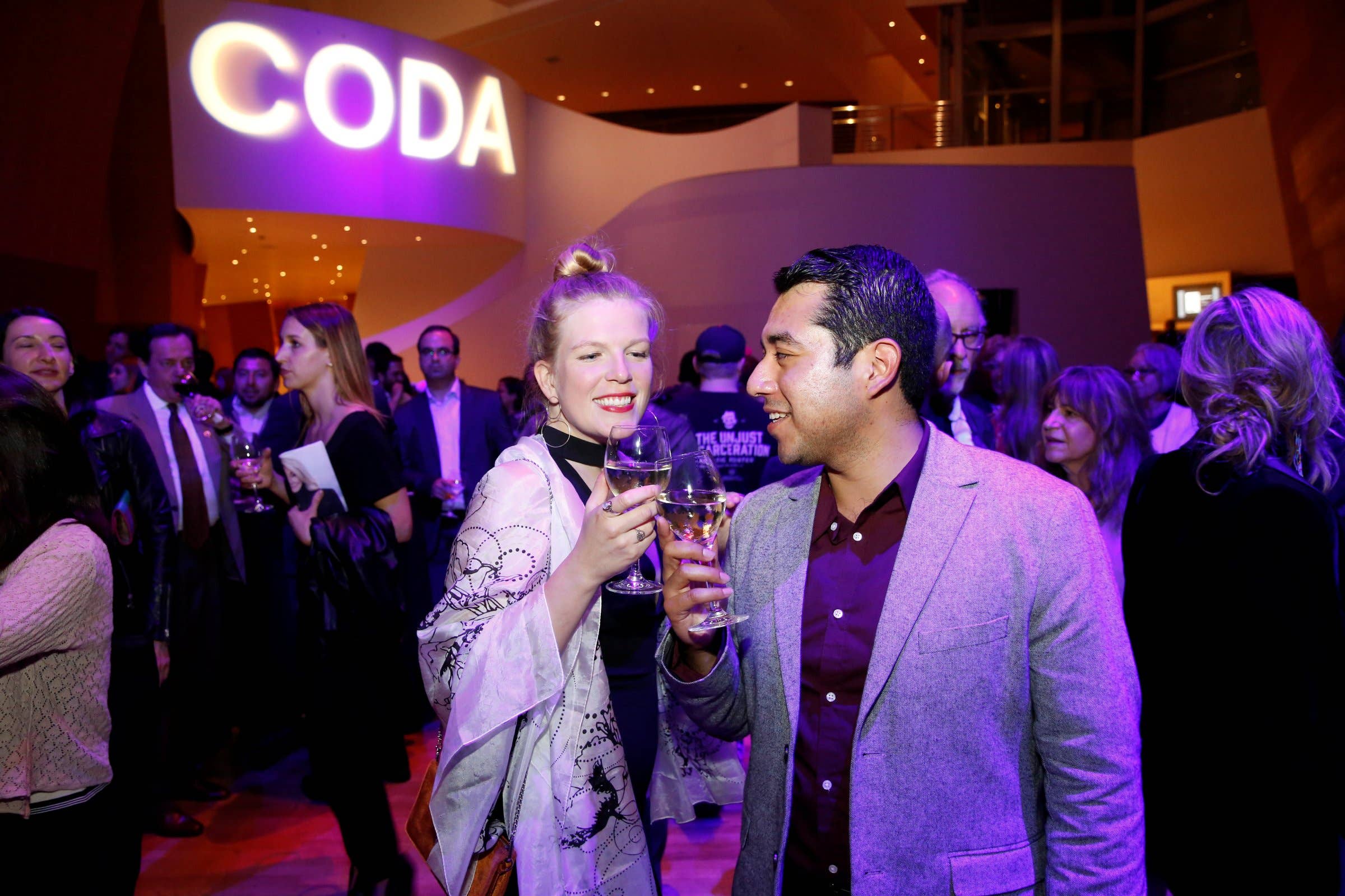 Guests toast with champagne at a gala event under purple lighting, with a “Coda” sign in the background.