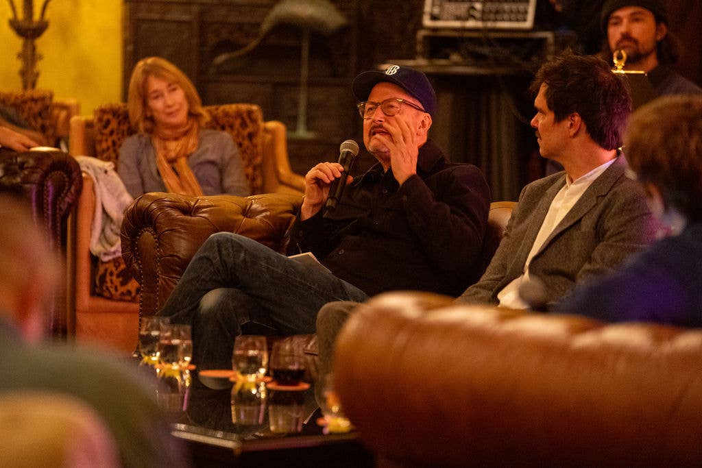 Man in cap holds a microphone at a panel discussion, seated on a couch with other people watching nearby.