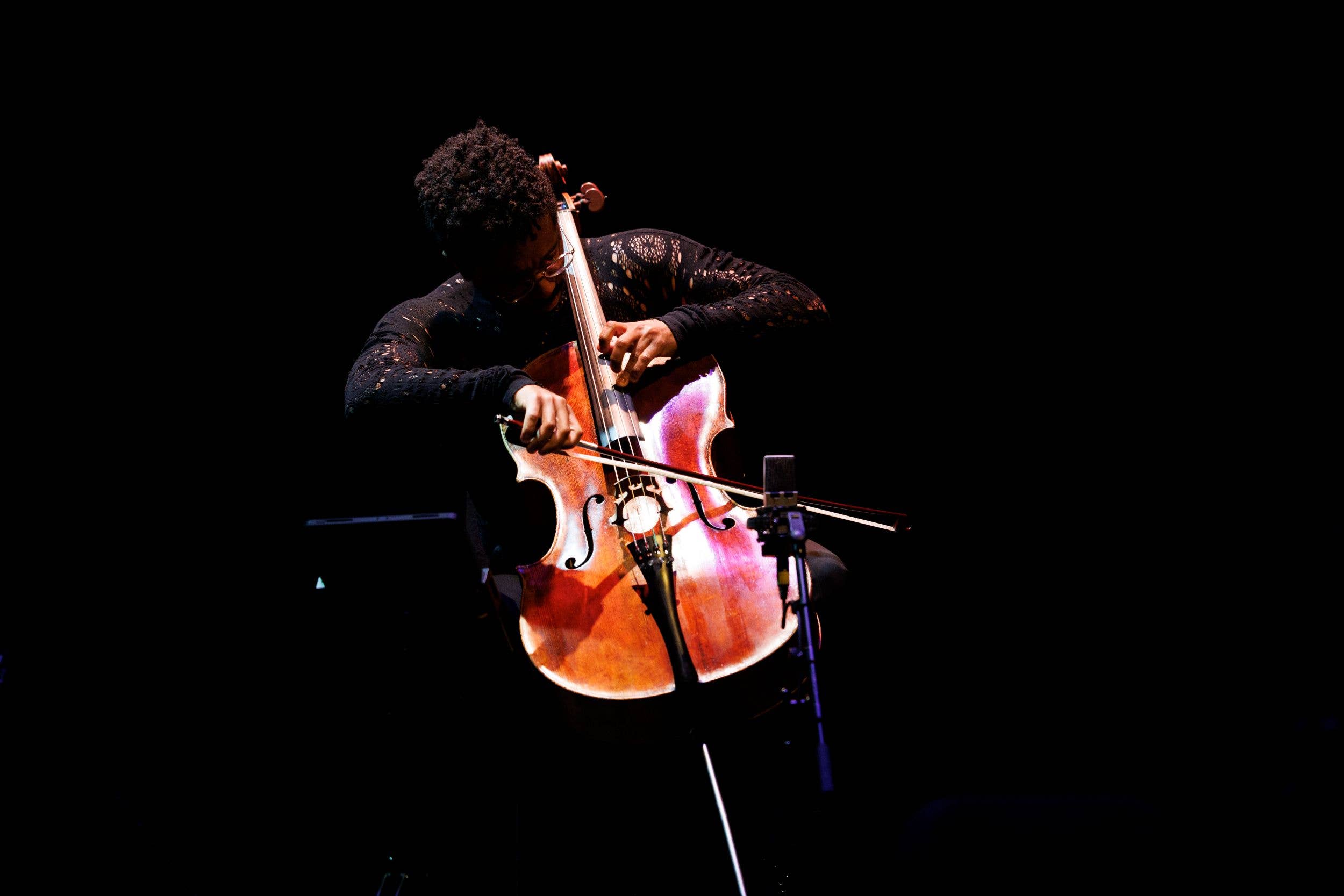 Musician in dark stage lighting playing an amber violin with a bow, standing behind a microphone.
