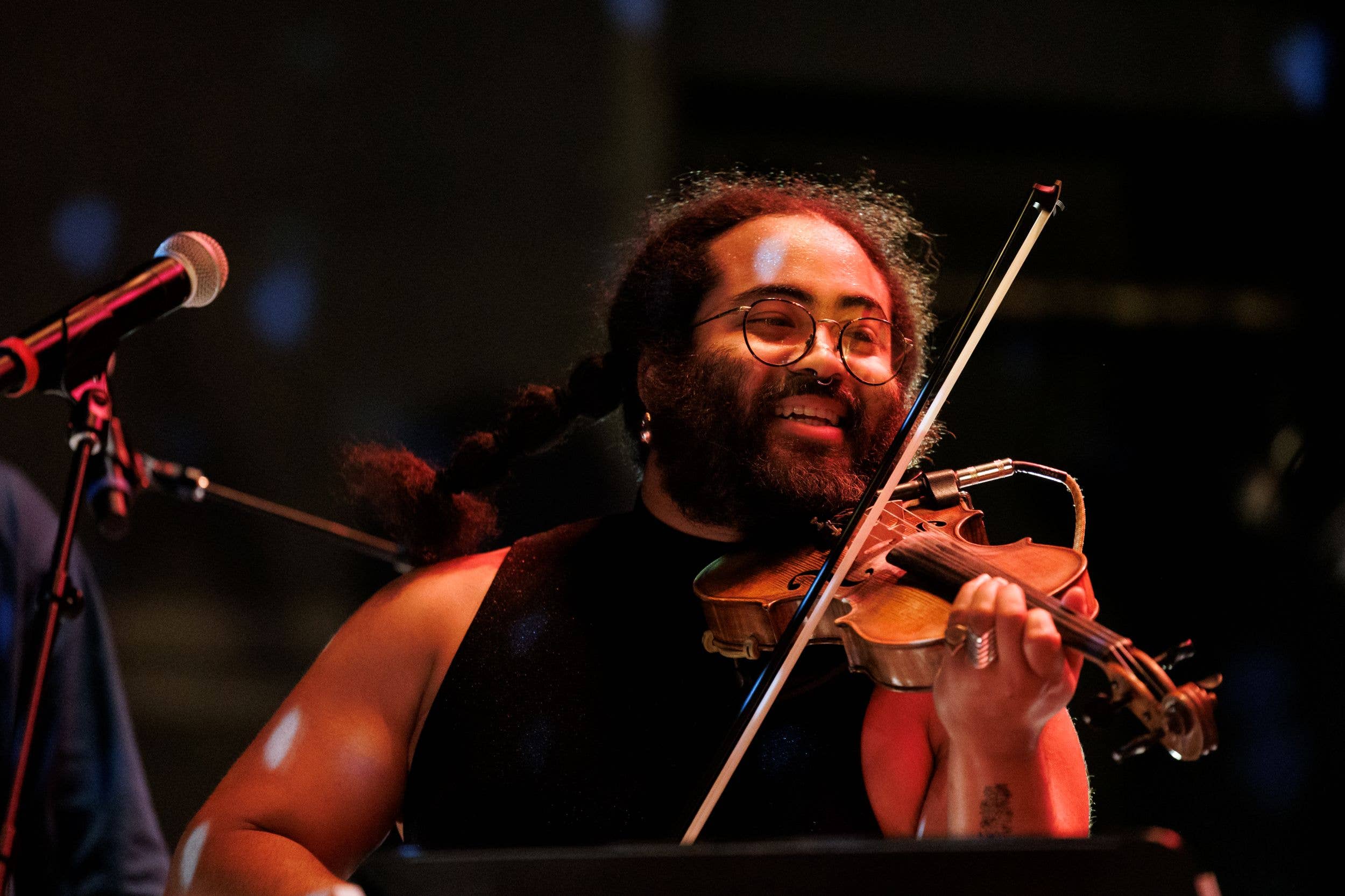 Violinist with curly hair and glasses performs on stage, holding a violin and bow near a microphone under warm lights.