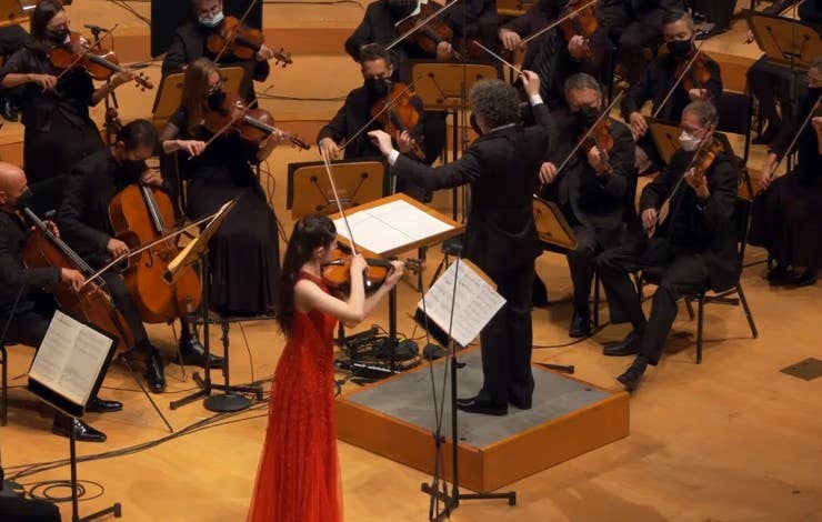 A woman in a red gown performs as an orchestra plays, with conductors and musicians in black masks and attire.