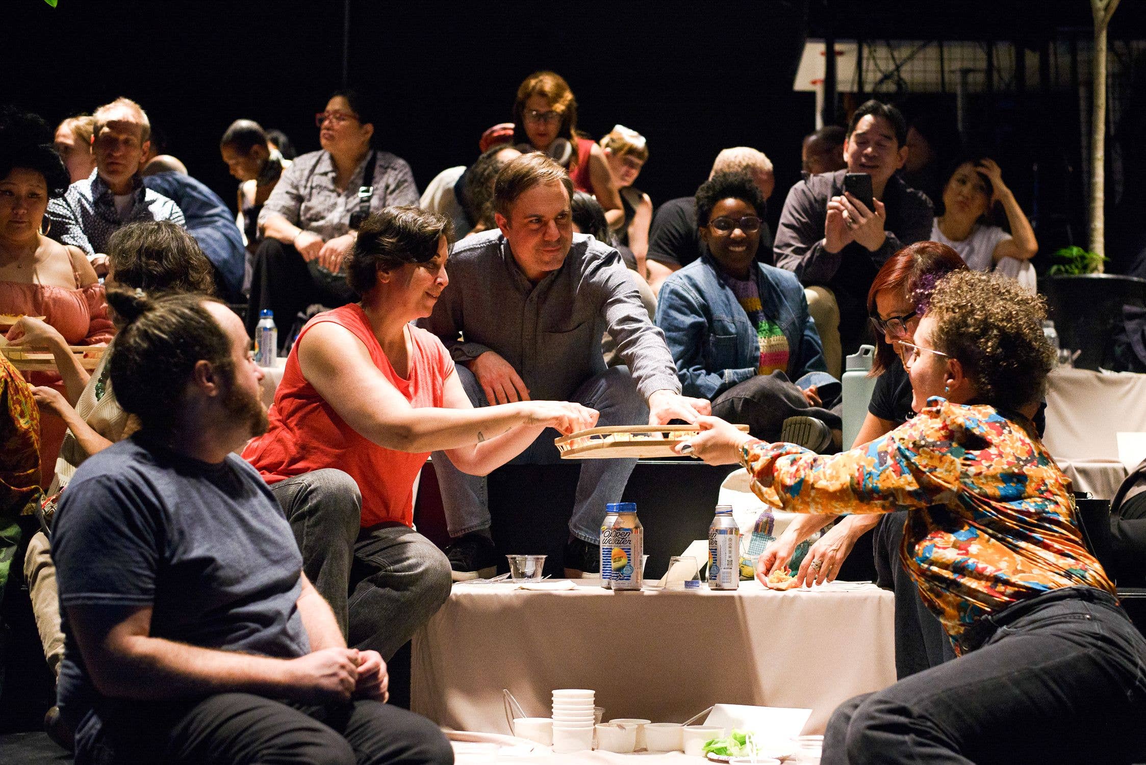 People seated at a convention table, smiling and reaching to share food and drinks under warm stage lighting