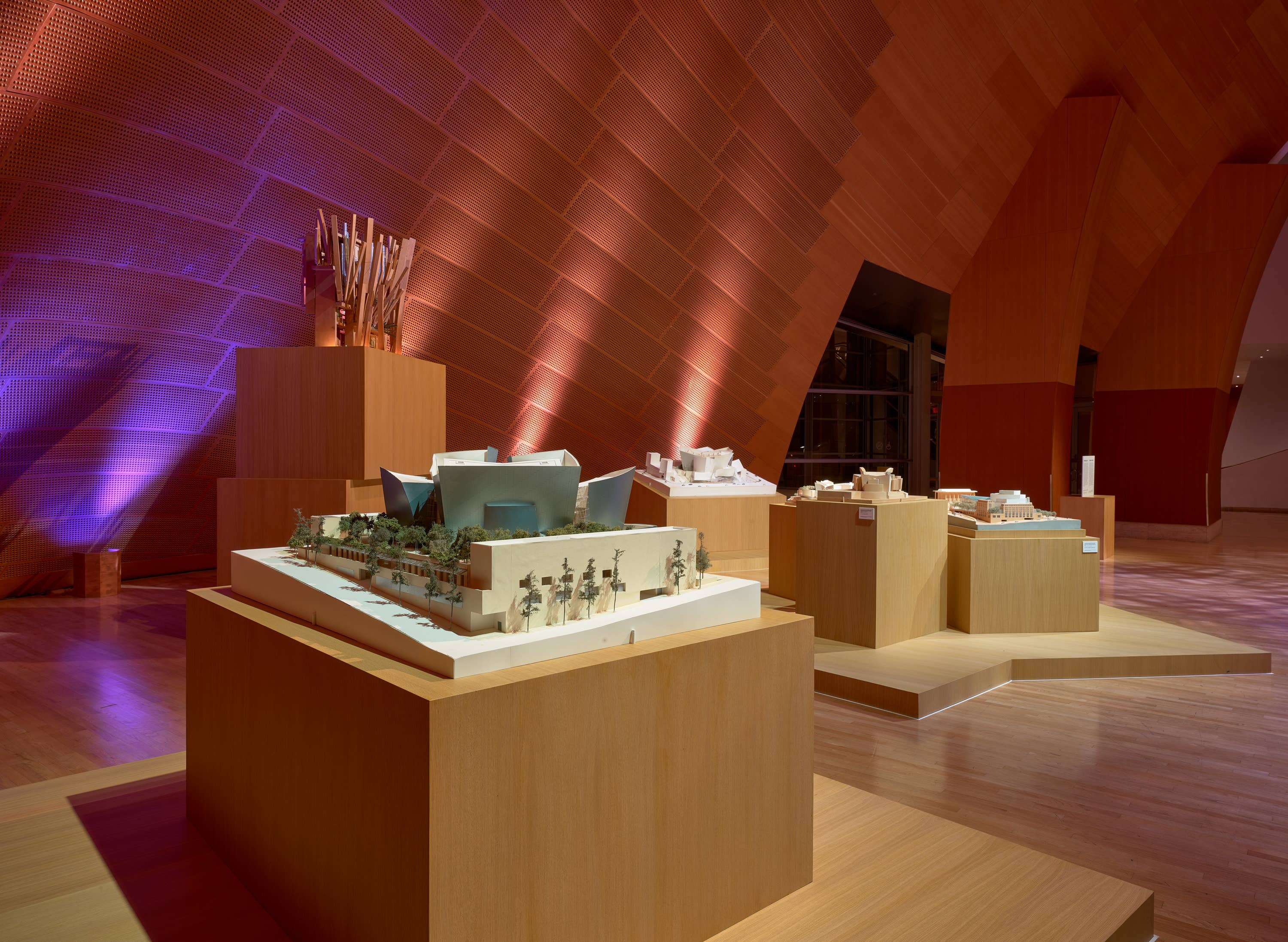 Architectural exhibition room with illuminated models and warm orange geometric walls and ceiling panels.