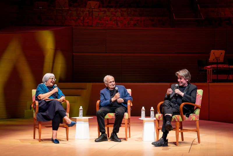 Three speakers sit on stage in orange chairs, holding microphones during a live panel discussion.