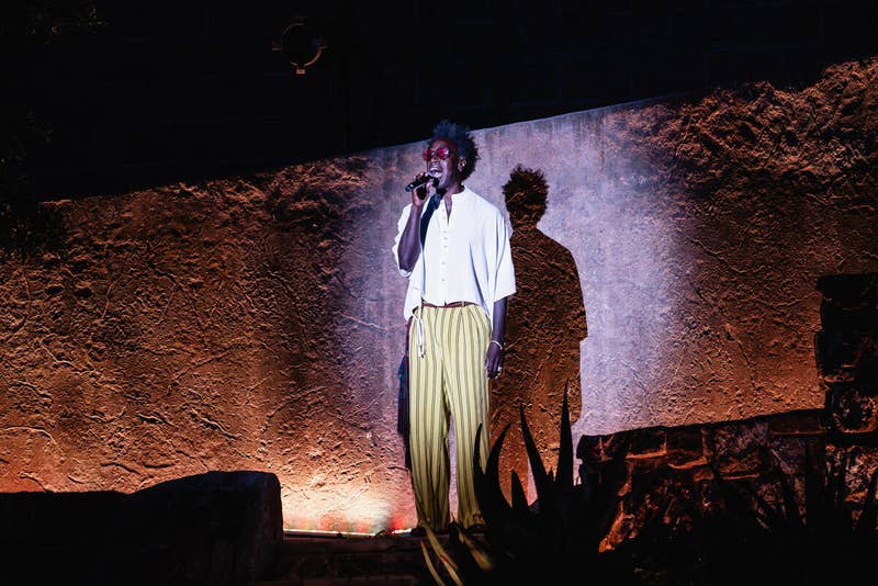 Singer in white jacket and striped trousers performs on stage under dramatic spotlight against textured backdrop.