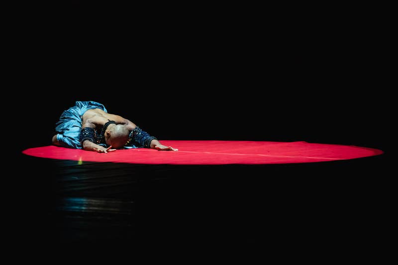 Two dancers in blue costumes lying on a red stage floor under dark lighting