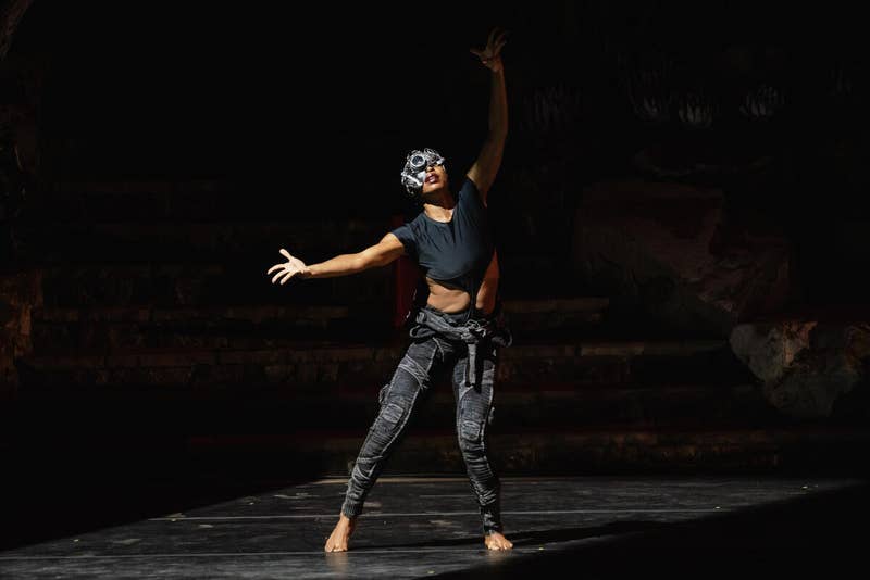 Performer in black and denim dances on a dark stage under a spotlight, arms raised and outstretched.