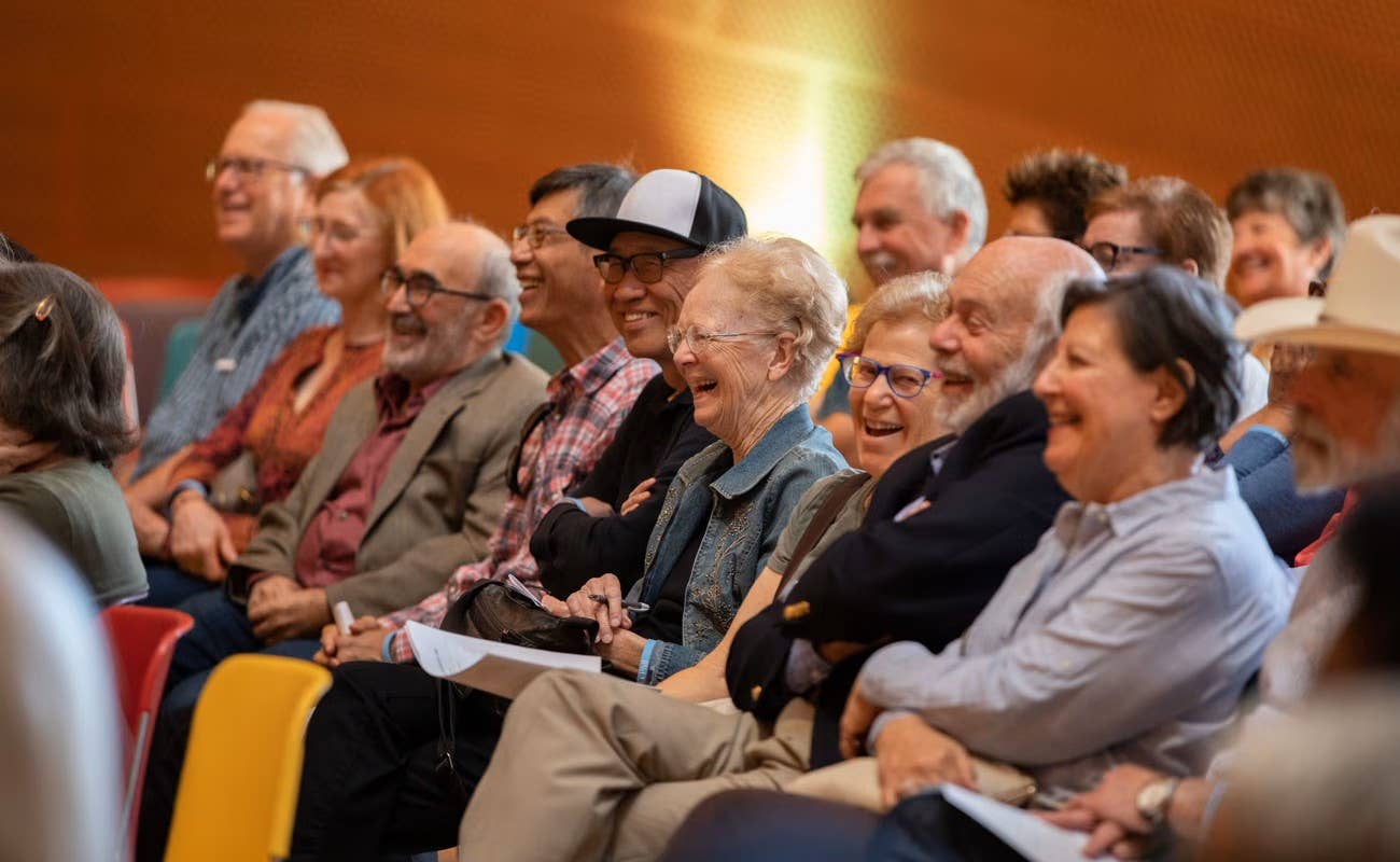 Audience of smiling seniors seated in a theater, listening to a talk under warm stage lights