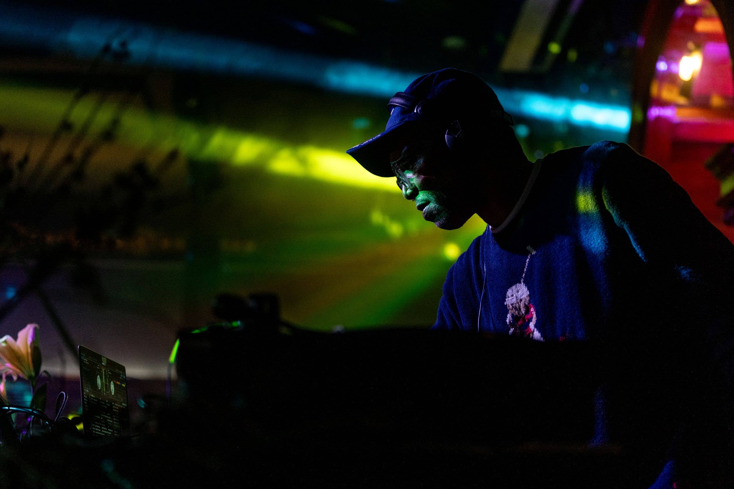 DJ wearing a cap and headphones, focused at a laptop with colorful neon lights in the background.