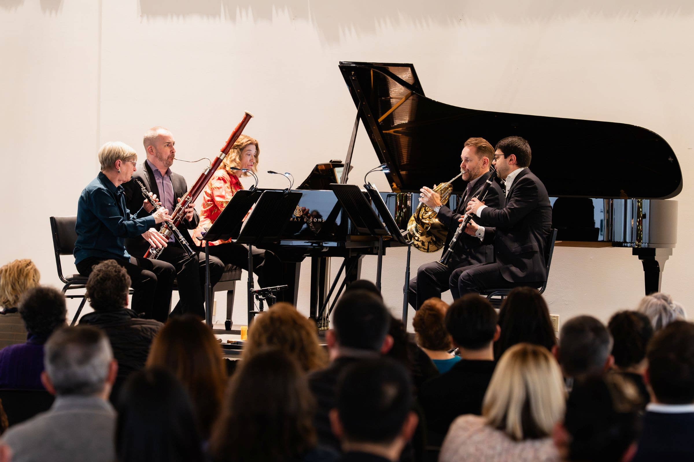 Orchestra musicians play clarinet, flute, and trumpet beside a grand piano at a concert, with audience in foreground