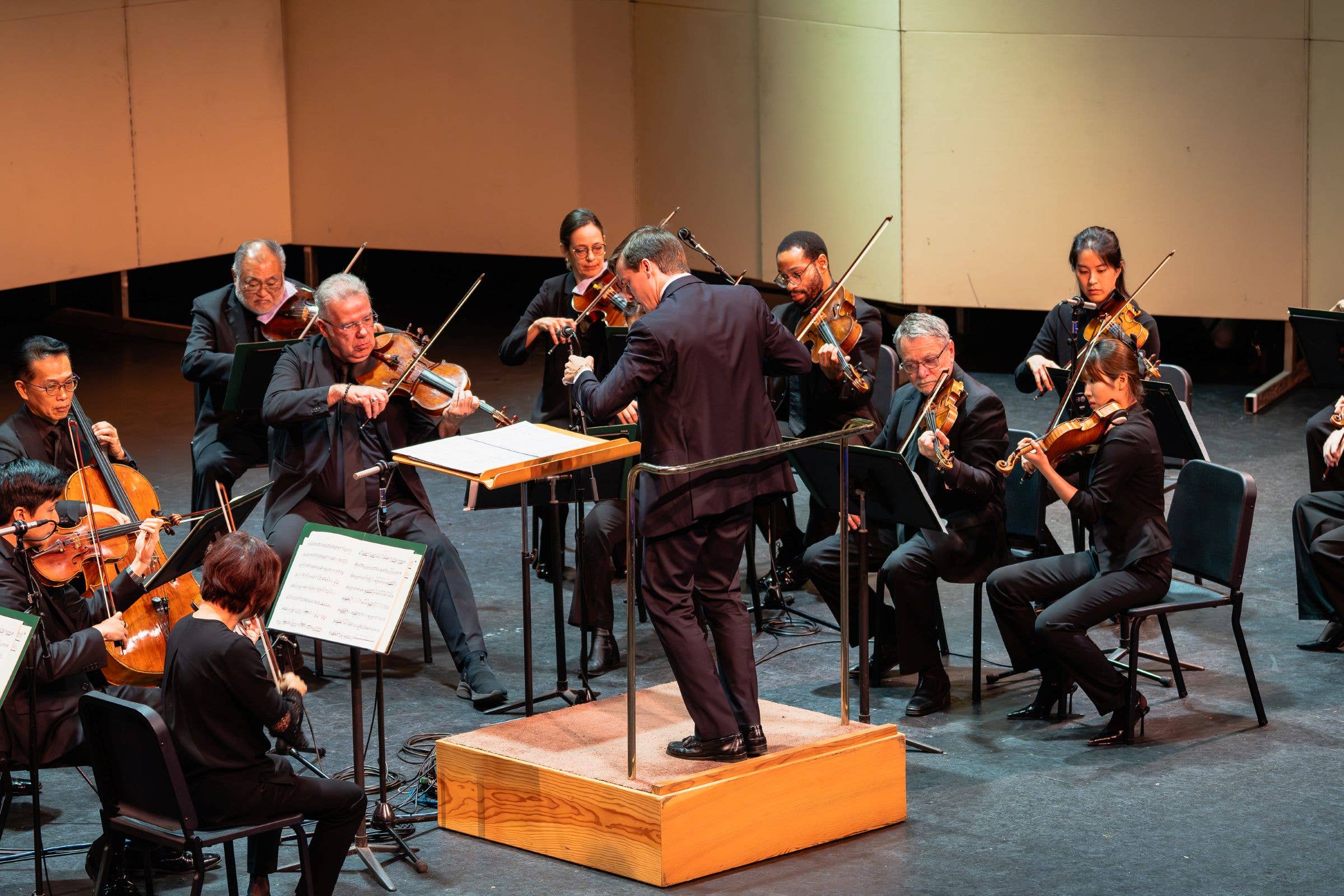 Orchestra string ensemble performing on stage with violinists and cellists under stage lights.