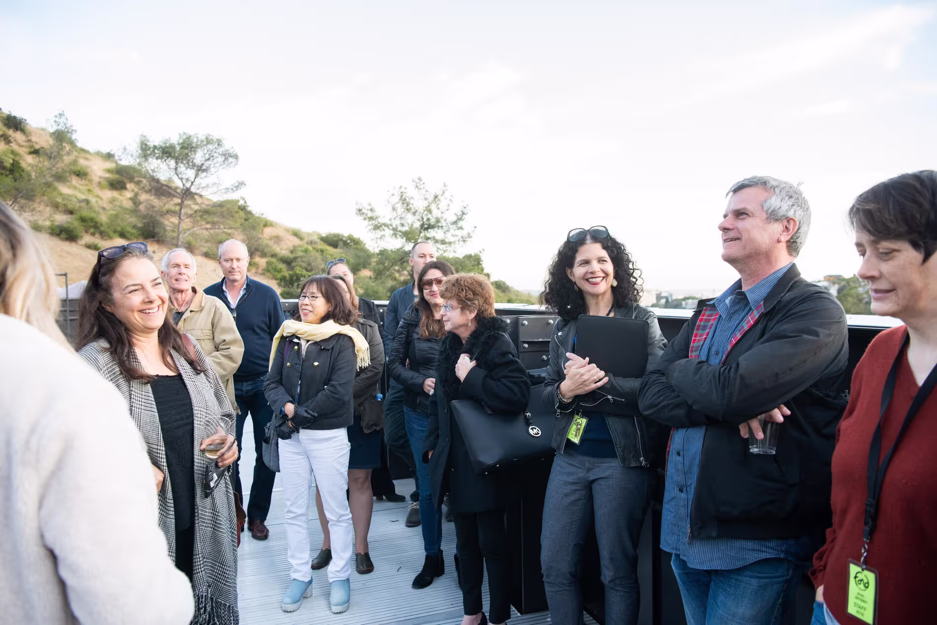 Group of people smiling and chatting on an outdoor bus, with hills and trees in the background