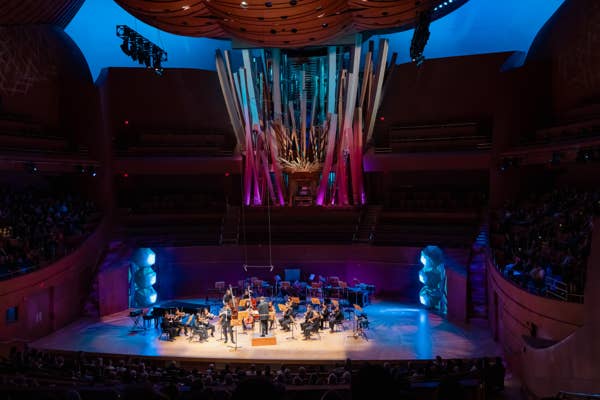Orchestra on stage beneath a glowing light installation, blue and purple stage lighting inside a concert hall.