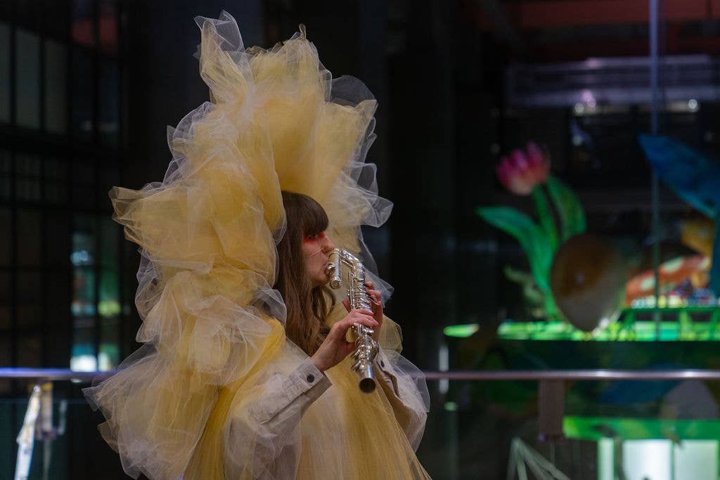 Woman in a flowing yellow tulle headdress sings into a microphone in front of colorful stage lights and instruments
