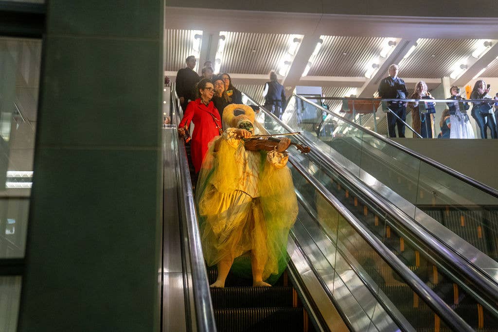 People ride an escalator in a modern indoor building, with a large costumed bear figure on the stairs.