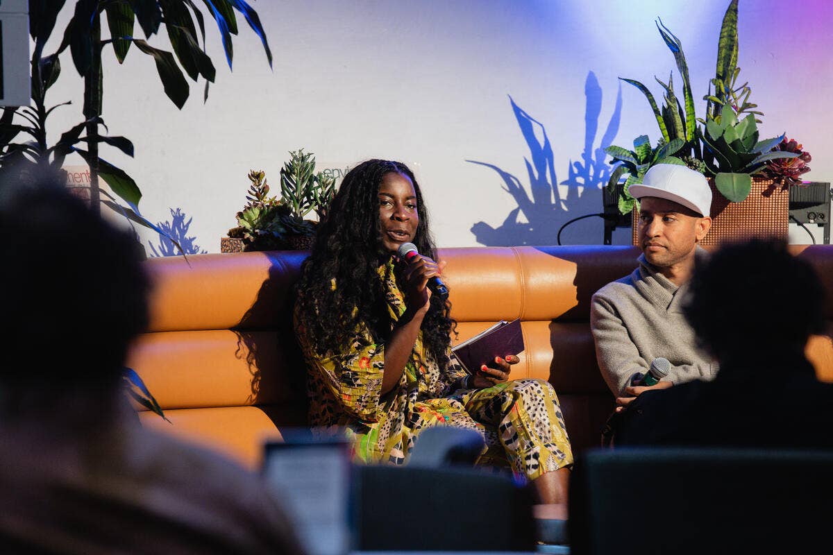 Woman in colorful outfit speaking into a microphone on an orange stage sofa, with a man in a cap listening.