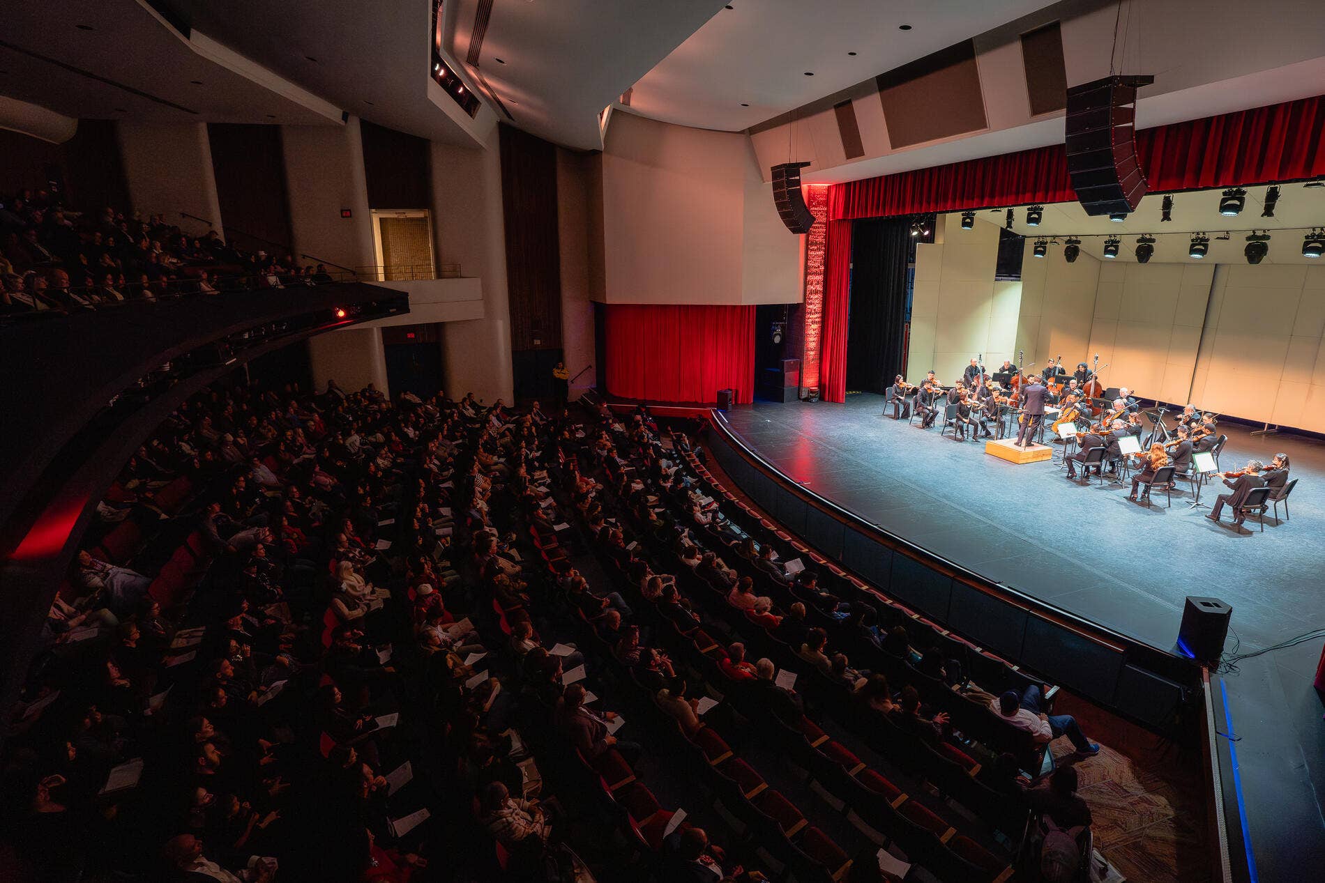 Concert hall with full orchestra on stage, red curtains, and an audience seated in tiered balconies.