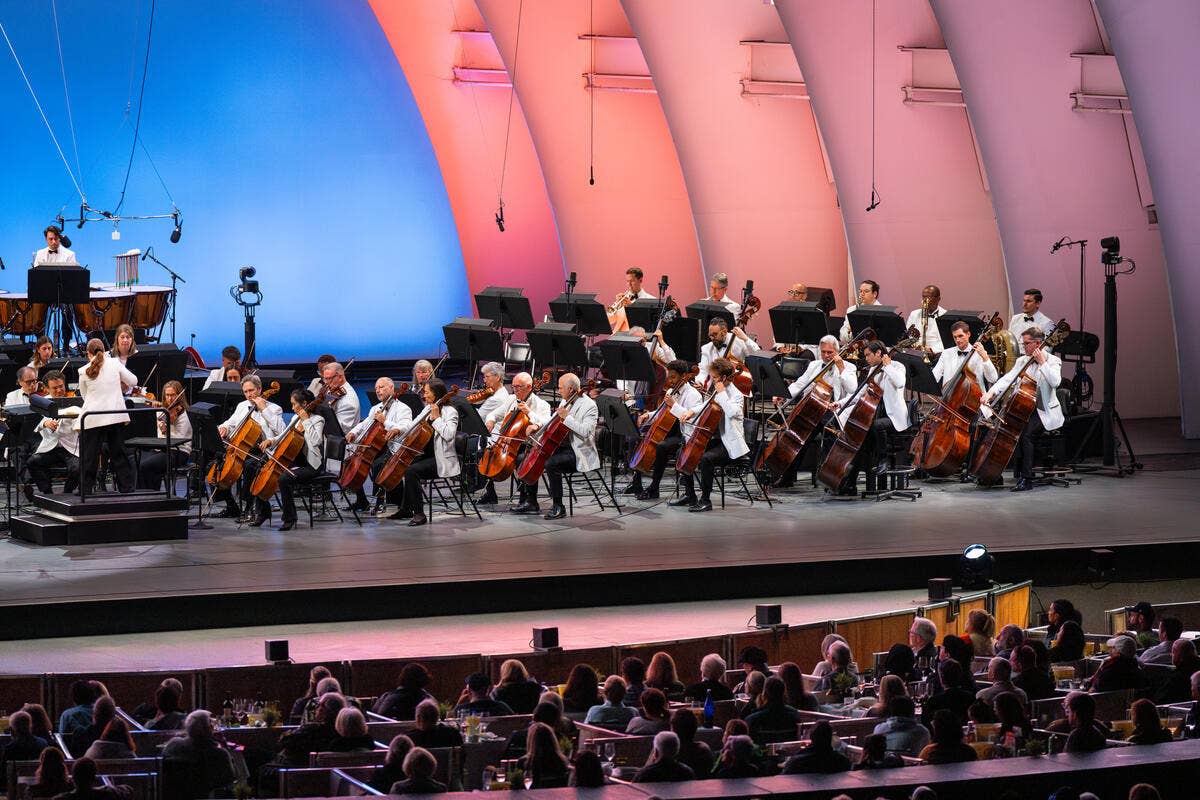 Orchestra seated on a concert stage with violins and brass under pink-lit architectural arches