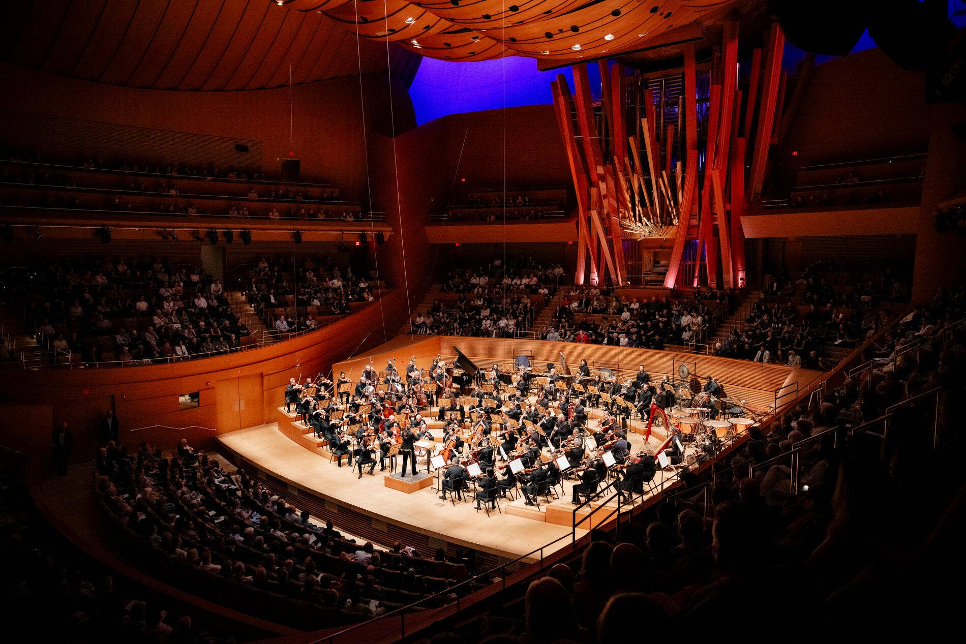 Orchestra performs on a lit stage in a concert hall with red framework and glowing blue backdrop
