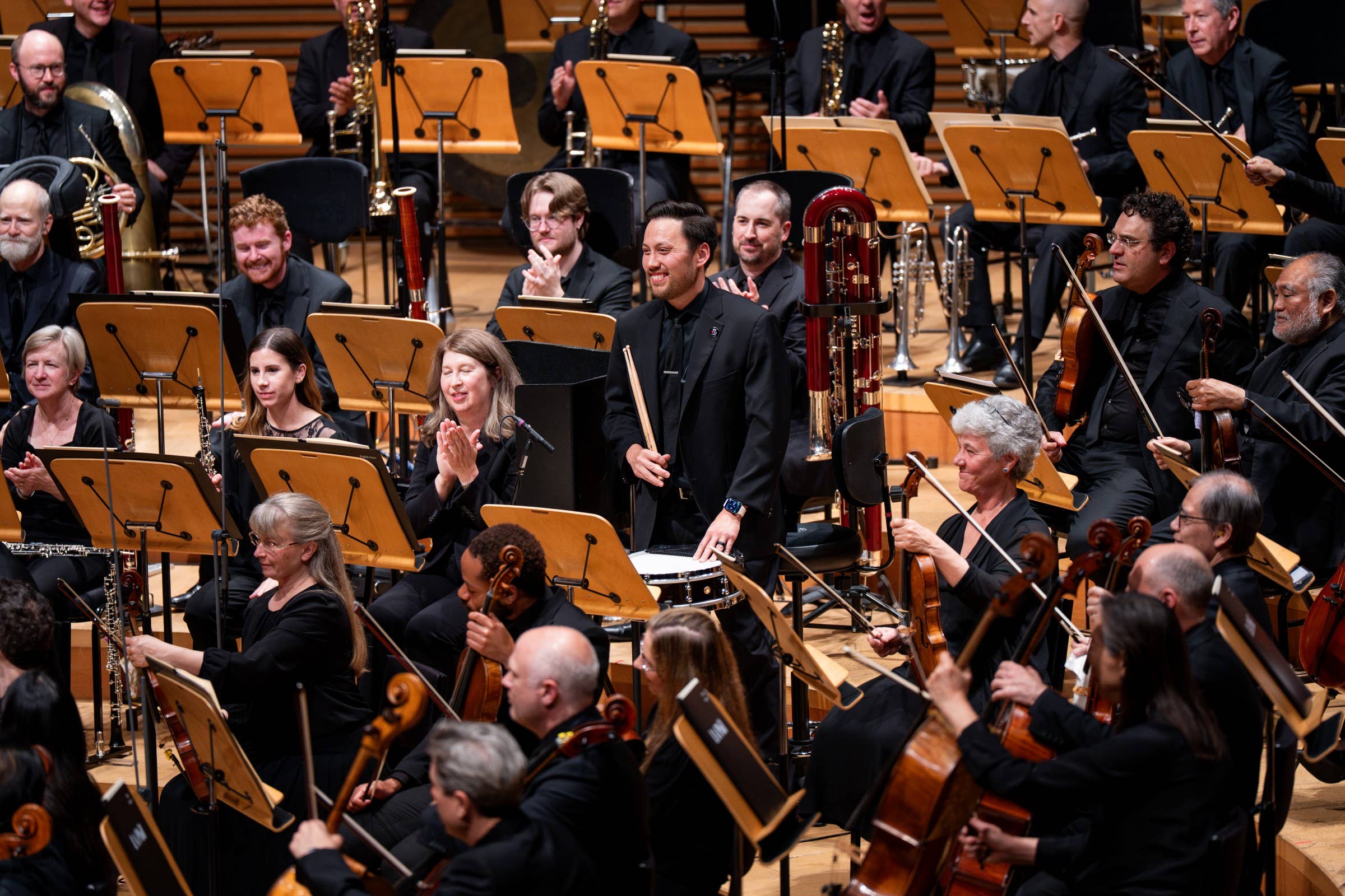 Orchestra seated on stage, musicians in black holding instruments, brass and saxophones in the front during performance.