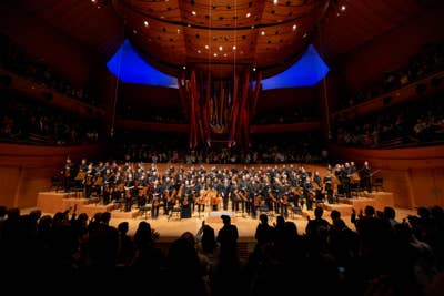 Symphony orchestra standing on stage with conductor, audience silhouettes cheering, surrounded by a curved concert hall ceiling