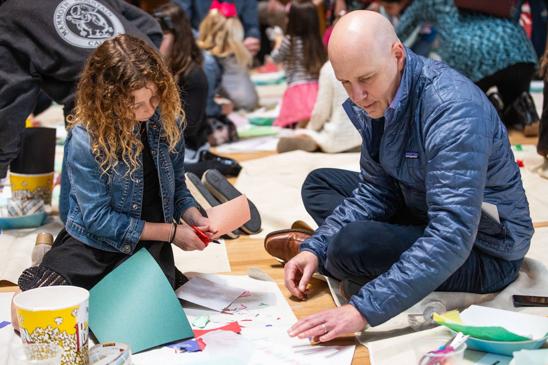 Two people sit cutting and assembling paper craft pieces during a community workshop.