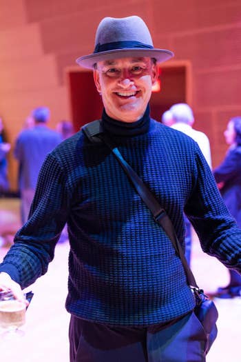 Smiling man in a blue hat and sweater gestures while holding a drink at a conference event.