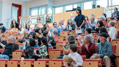 Audience members applaud in a lecture hall as a person stands at the front holding papers