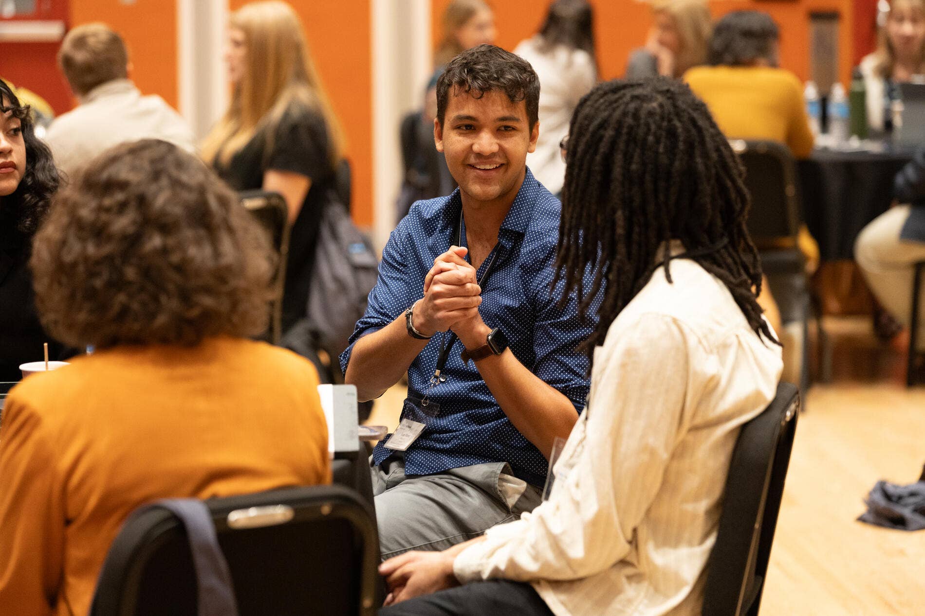 Man in a blue shirt smiles while conversing in a conference setting, seated at tables with attendees around him.