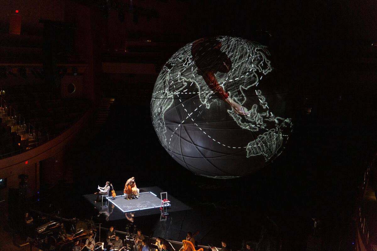 Stage performers under a hovering glowing globe with light routes and a red path in a dark theater.