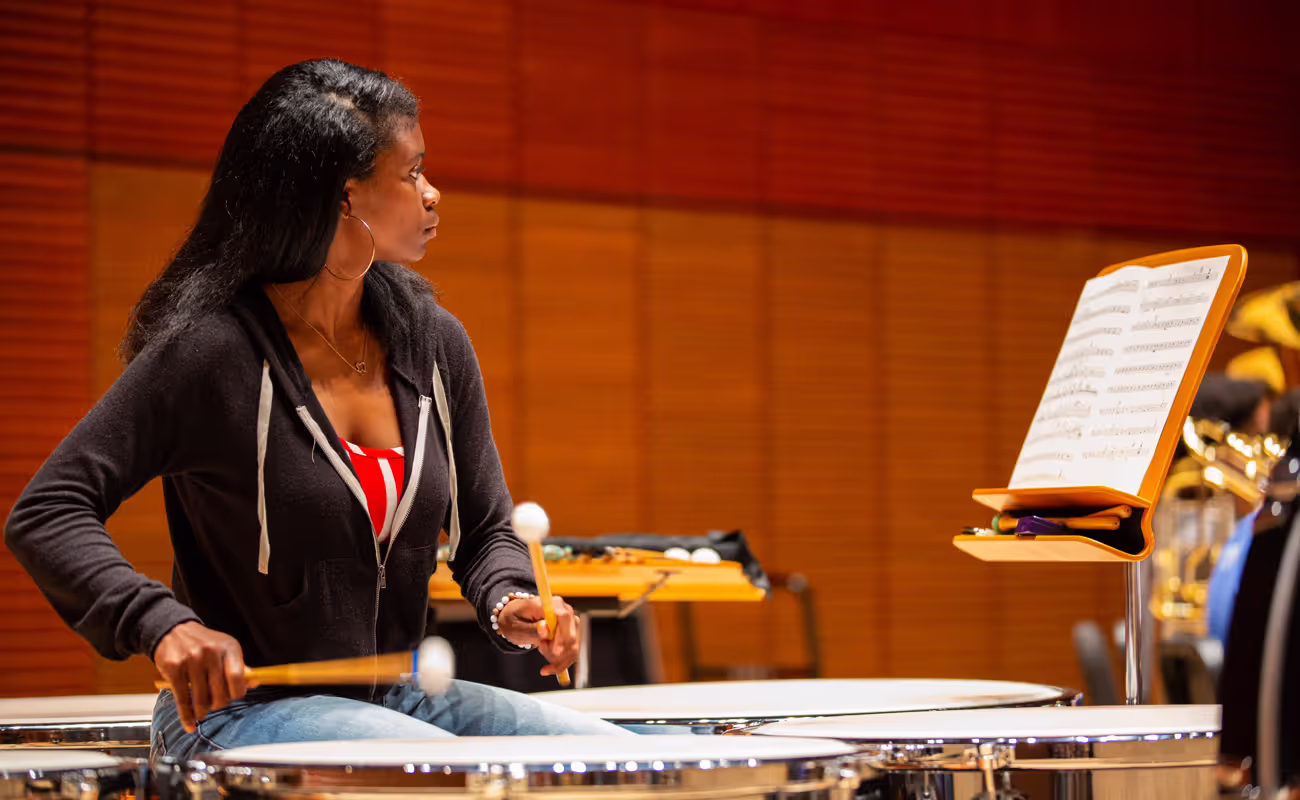 Person plays timpani while reading sheet music on a stage.