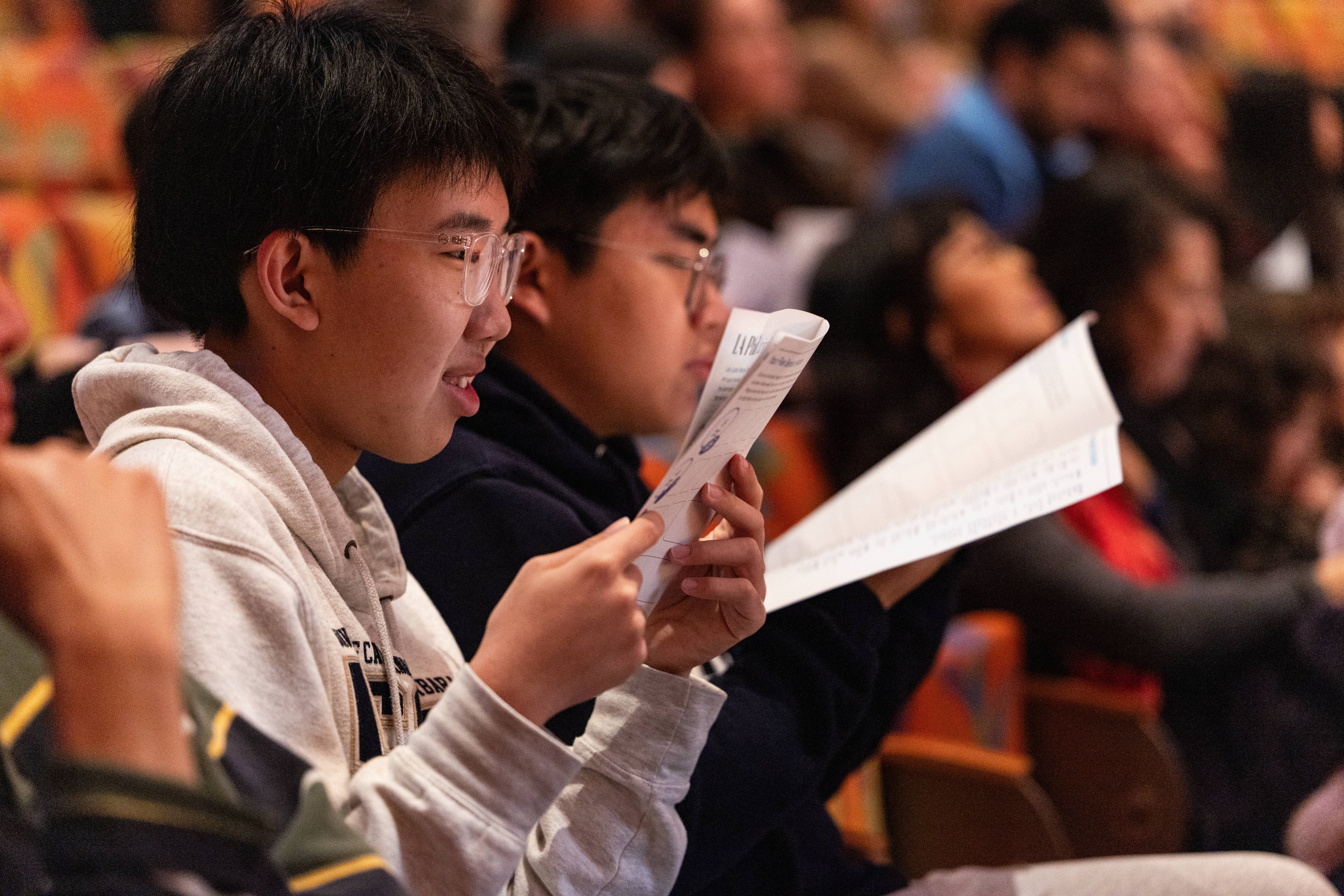 Students wearing glasses read printed worksheets in a classroom with colorful, blurred background lights.